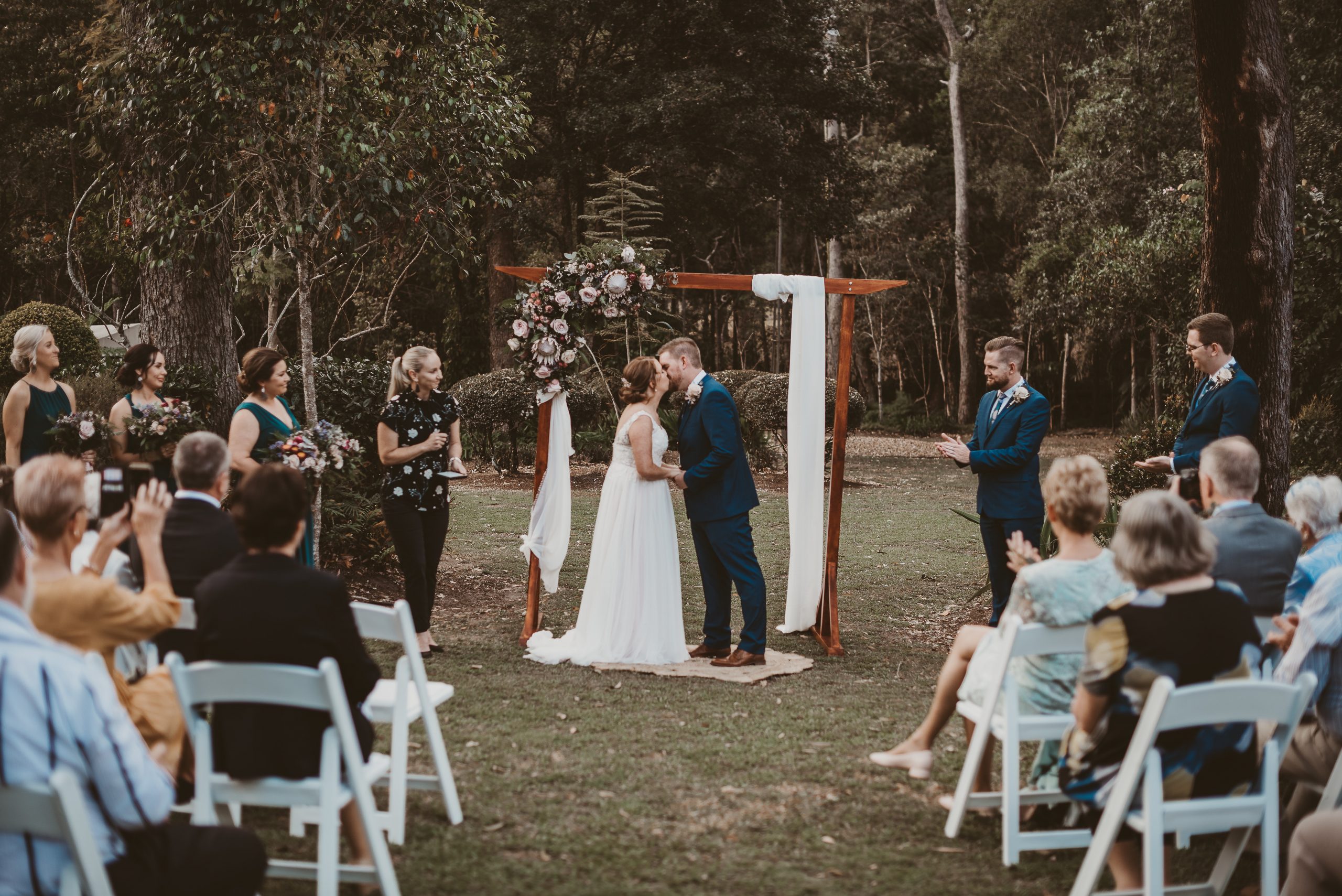 A couple kiss at their ceremony in the gardens, guests seated either side