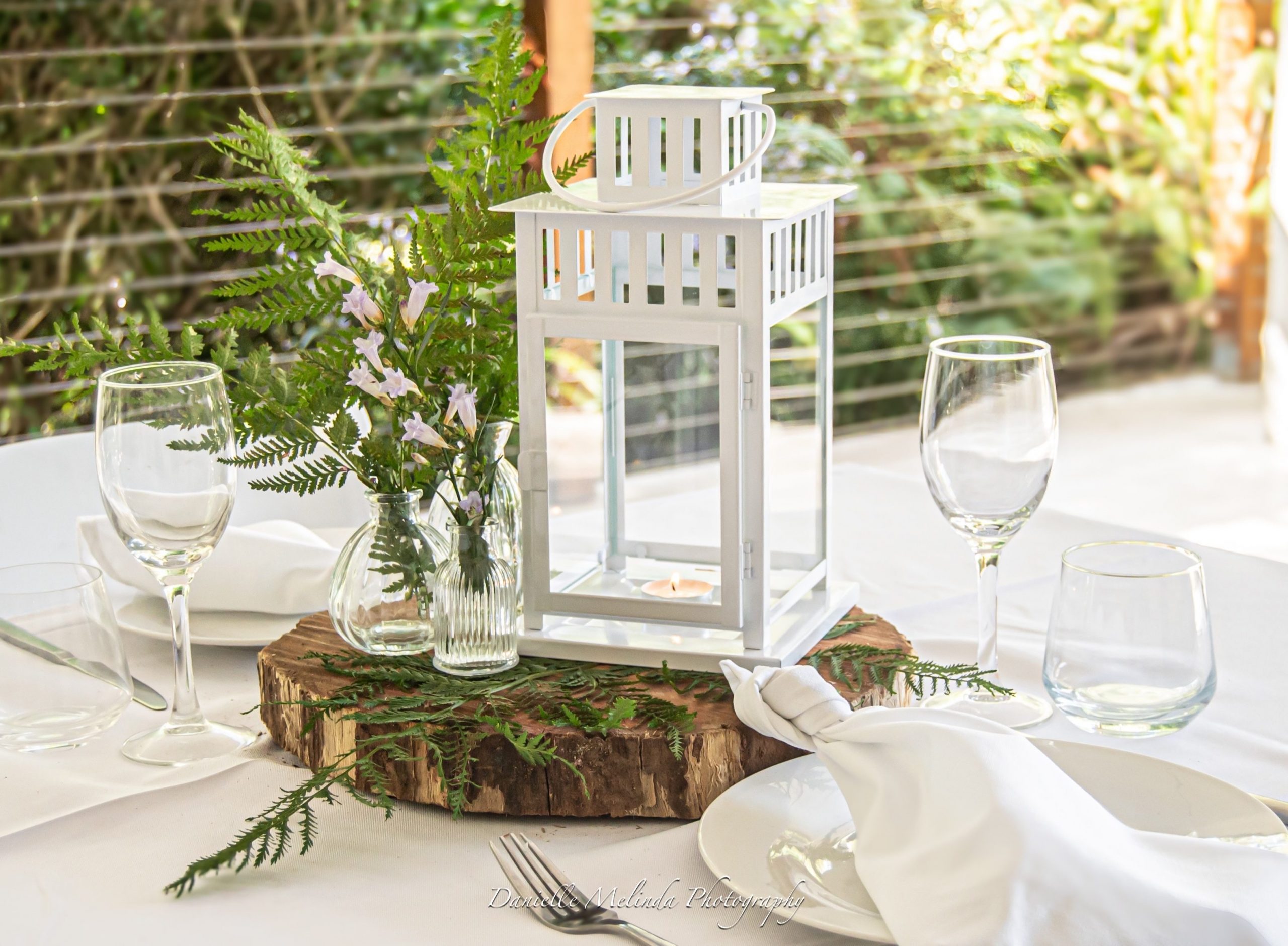 Table centrepiece with a white lantern, ferns and bud vases on a timber round